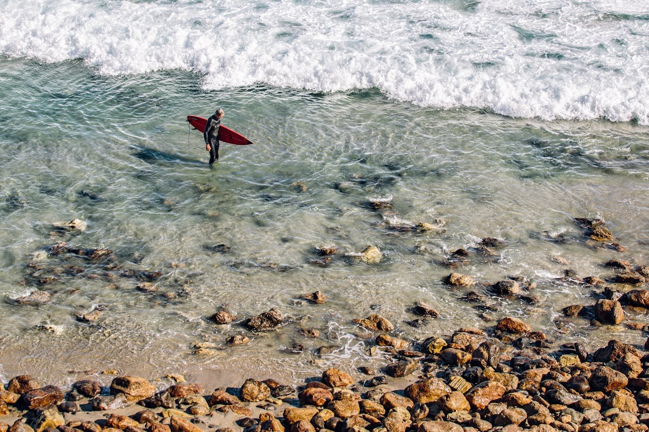 A lone surfer carrying a red surfboard walks along a rocky shoreline with gentle ocean waves.