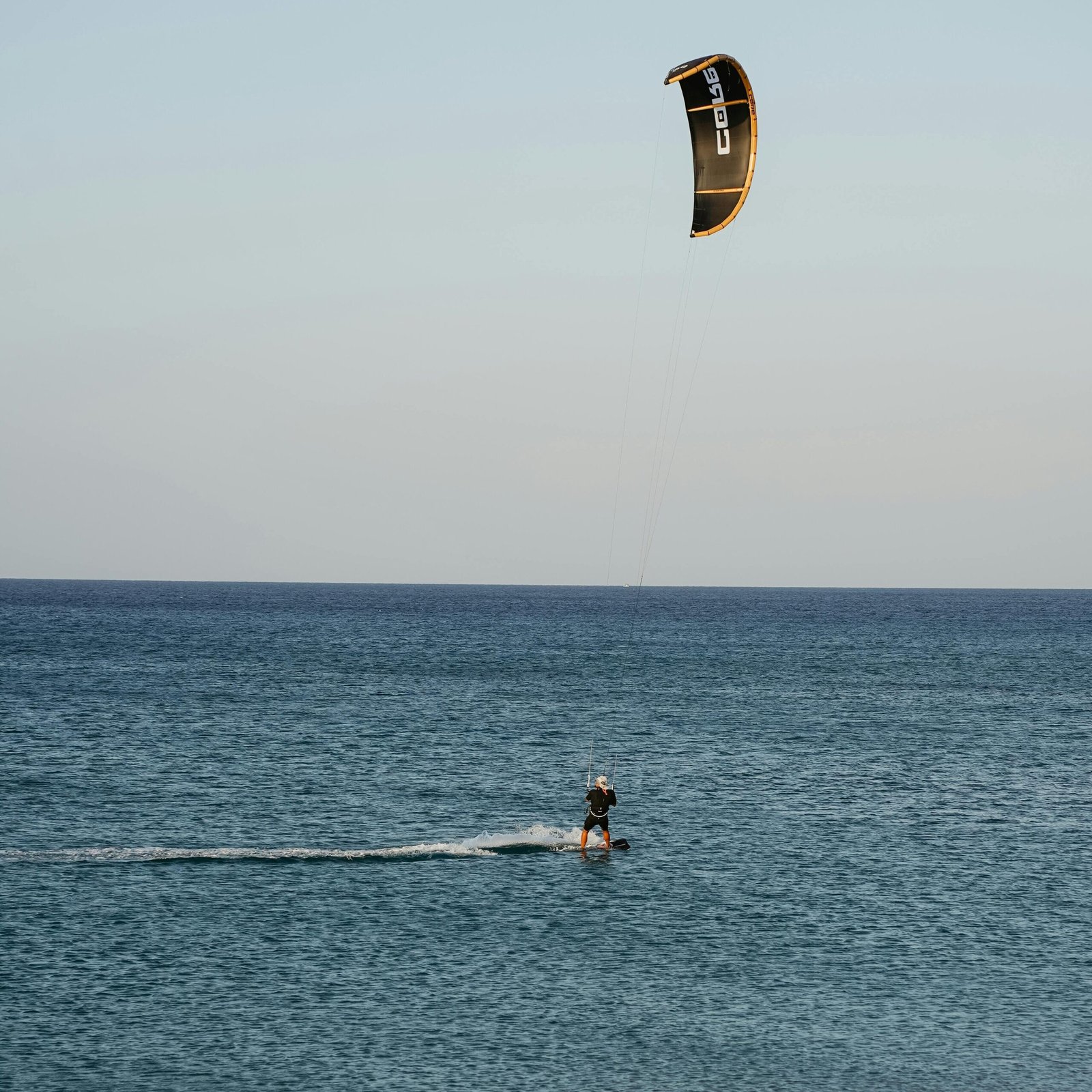 Kitesurfer gliding over calm ocean waves under a clear blue sky at sunset.